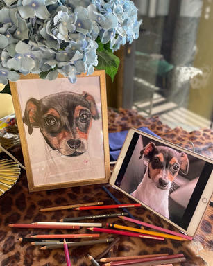 A finished framed portrait of a black-and-tan dog standing beside the tablet showing the photograph it was drawn from, with coloured pencils scattered across a leopard-print cloth.